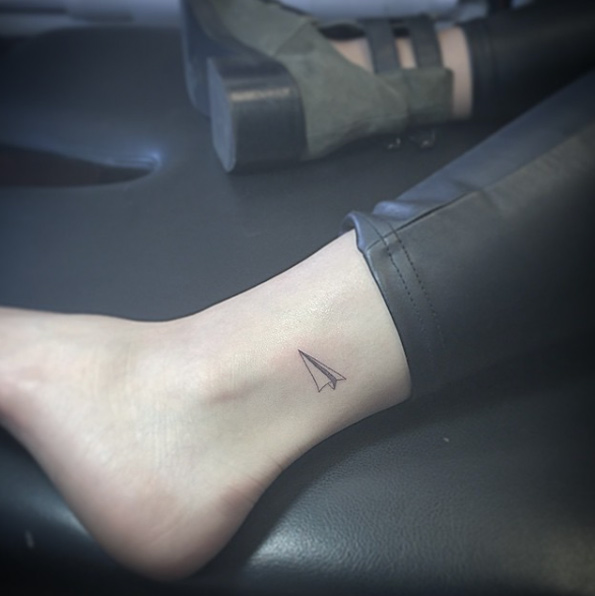 Tiny paper airplane tattoo on ankle by East Iz
